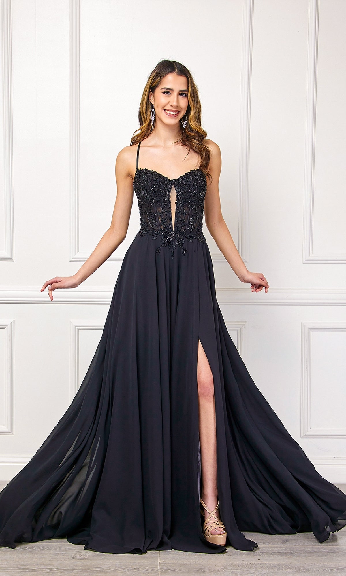 A woman in the Amelia Couture 7066 long black corset prom dress stands against a white paneled wall, smiling with her arms slightly extended.